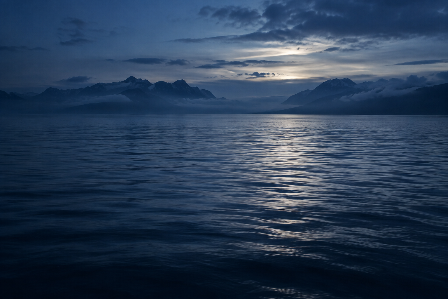 Silver light falls across a still blue sea, with a mountainous coast fading into mist.